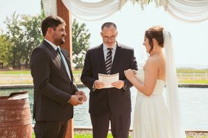 A wedding ceremony takes place outdoors on a sunny day with white curtains and beautiful willow trees in the background.