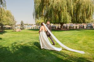 A flowing white wedding dress train trails across green grass beneath weeping willow trees.