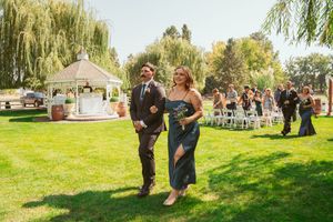 A couple walks down the aisle after their outdoor ceremony near a white gazebo and willow trees.