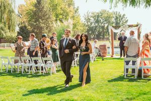 People walk in a line outdoors at a scenic garden venue with chairs set up on green grass during a sunny day.