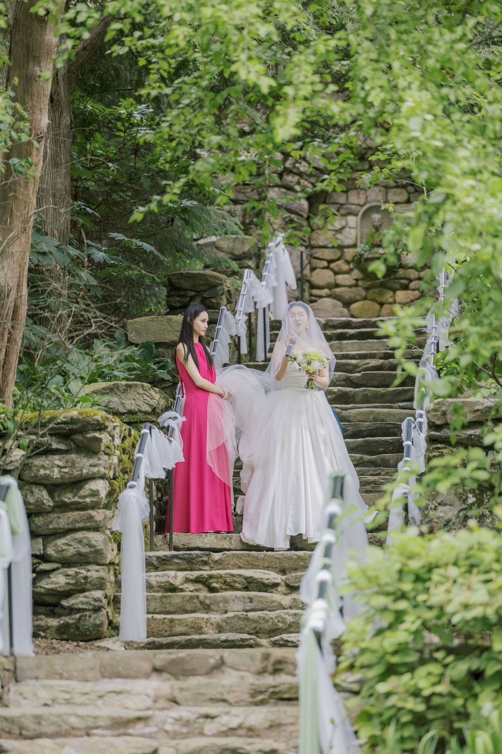 Stone staircase adorned with white decorations leads through lush greenery at an outdoor wedding venue.