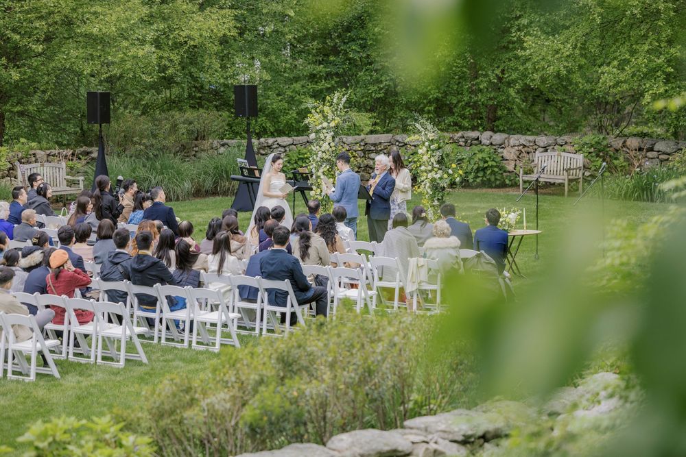 Outdoor wedding ceremony setup with white chairs and floral arch surrounded by natural greenery.