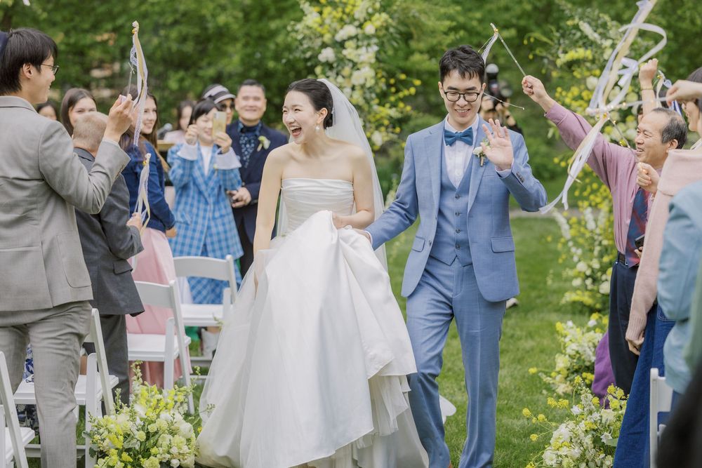 Wedding guests celebrate outdoors by blowing bubbles as a newlywed couple walks down an aisle lined with flowers.