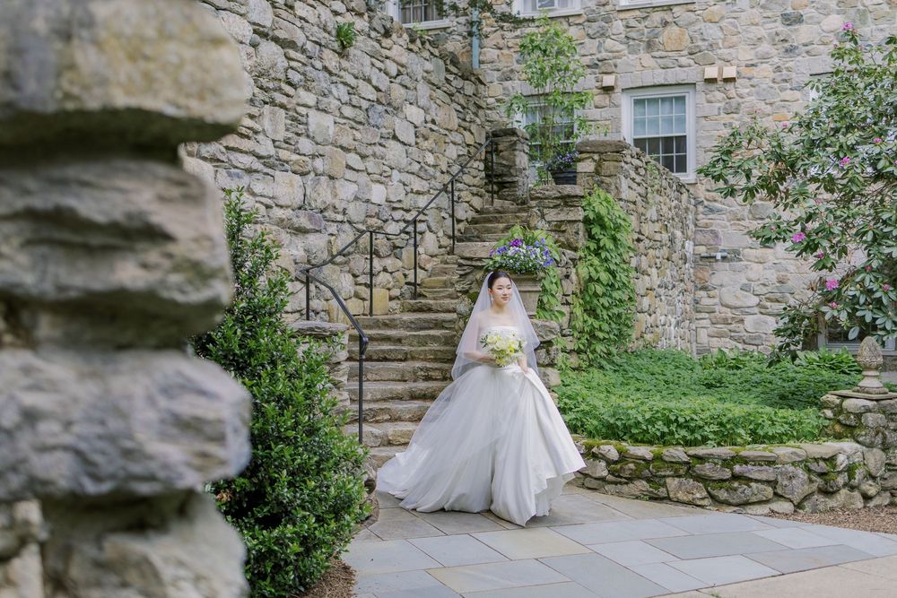 Bride in flowing white gown ascending stone staircase at rustic outdoor venue.