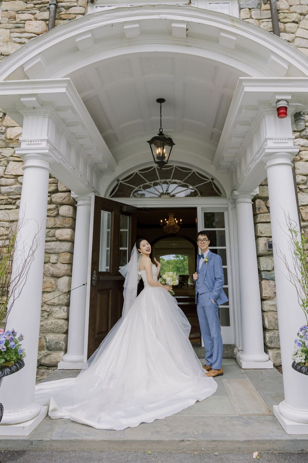 A wedding couple stands in an elegant stone archway entrance with white columns and impressive architectural details.