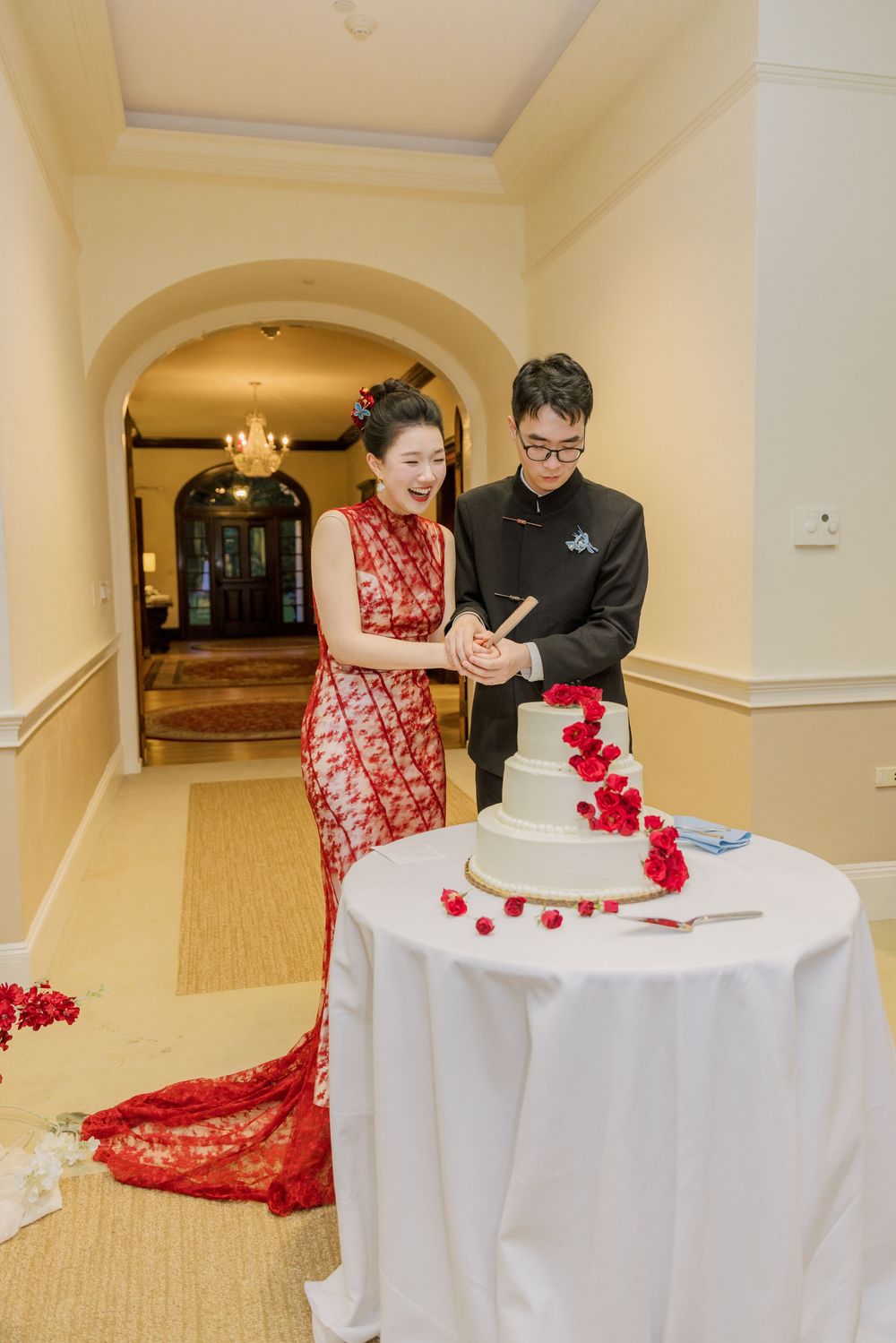 A couple cuts their white wedding cake decorated with red flowers at an indoor reception.