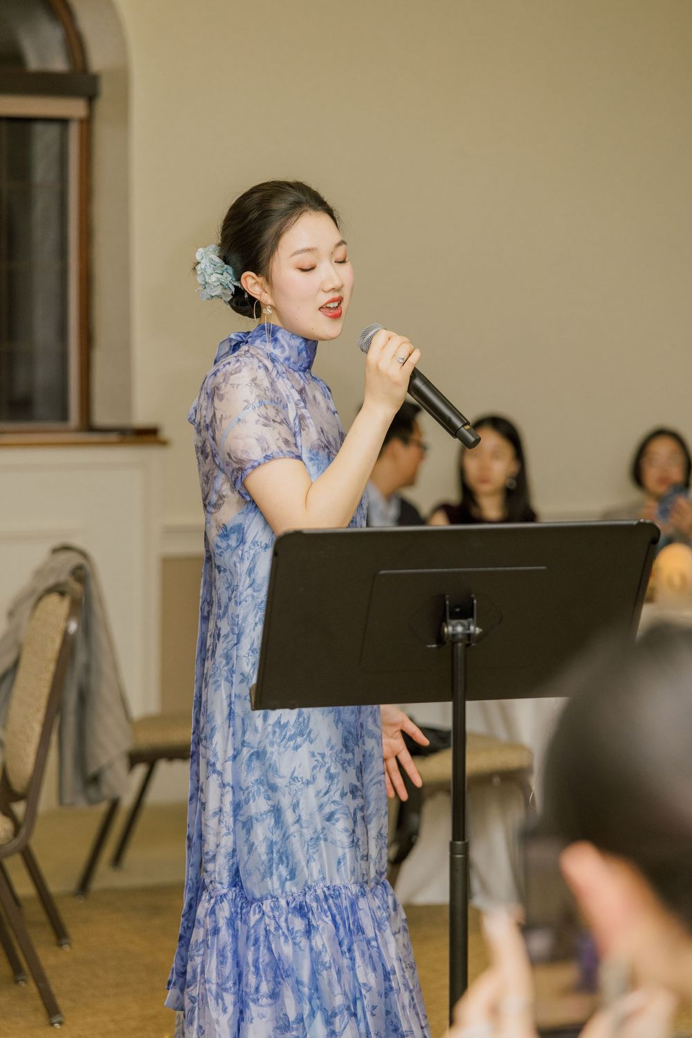 A performer in a blue floral dress sings at a music stand during an indoor event.