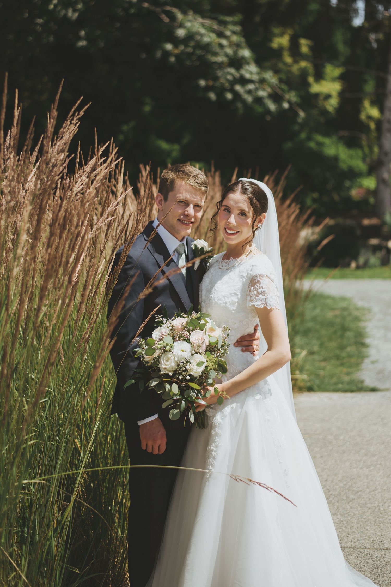 A newlywed couple stands together by ornamental grasses during an outdoor garden wedding photoshoot.