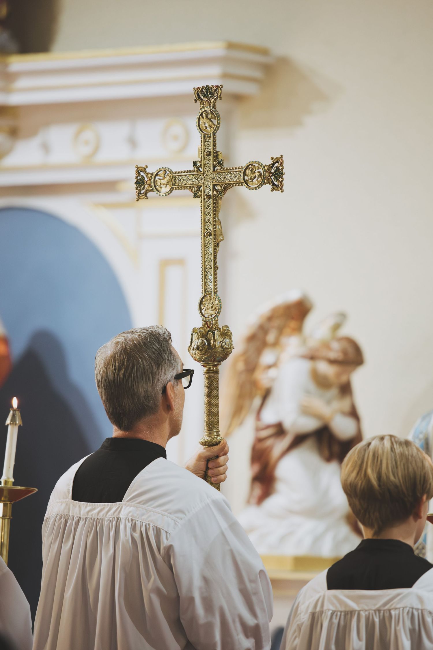 Religious figures in white robes holding an ornate golden cross during a church ceremony with decorative wall artwork.