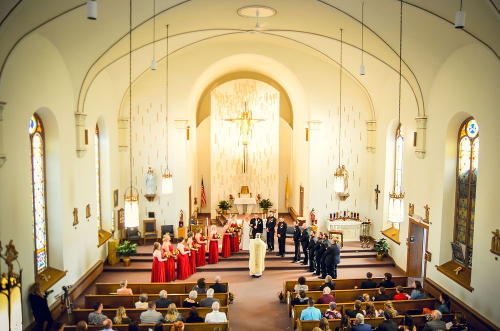 A church wedding ceremony takes place in a traditional white chapel with red-robed clergy and guests in the pews.