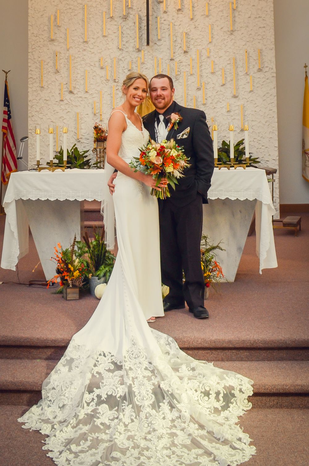 Elegant wedding couple poses near church altar with white lace train dress and colorful floral arrangements.