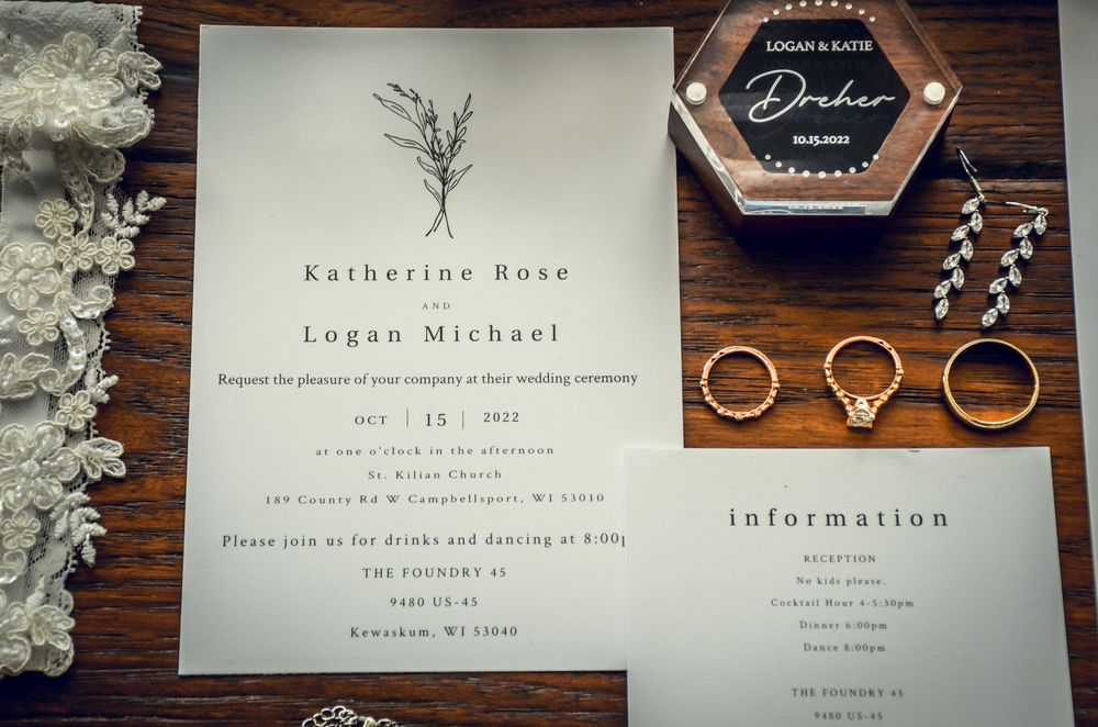 Wedding invitation suite displayed on dark wooden surface with floral accents and wedding rings.