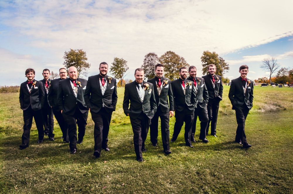 Wedding party in black tuxedos and red ties gather on a grassy hill under blue sky during autumn ceremony.