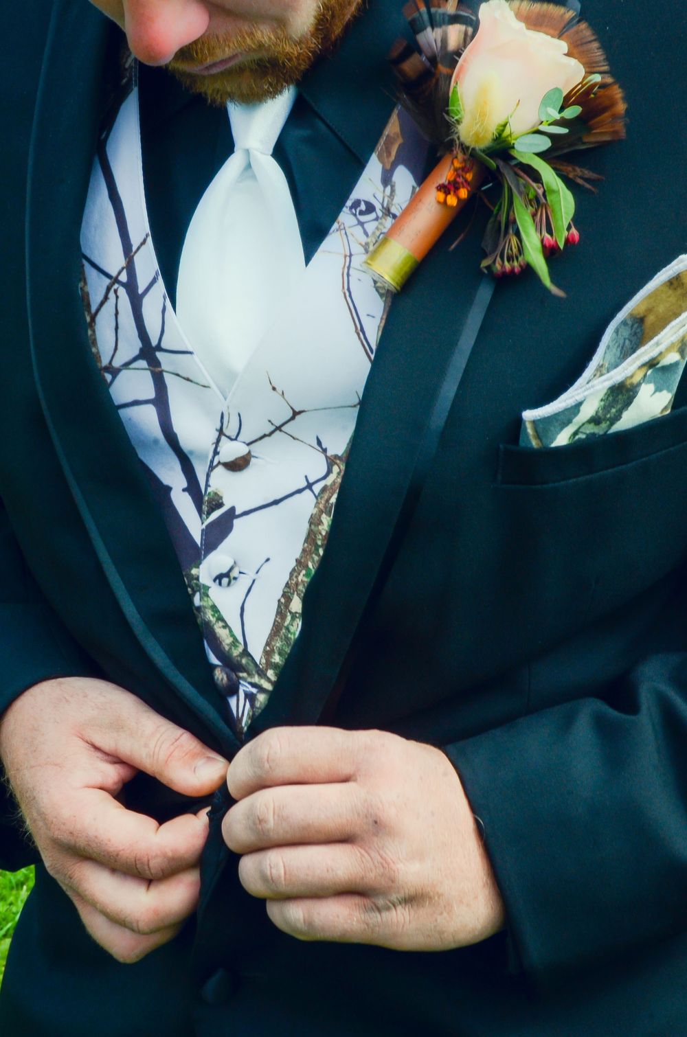 Close-up of groom's dark suit jacket with white camouflage vest and white tie, featuring a rustic boutonniere.
