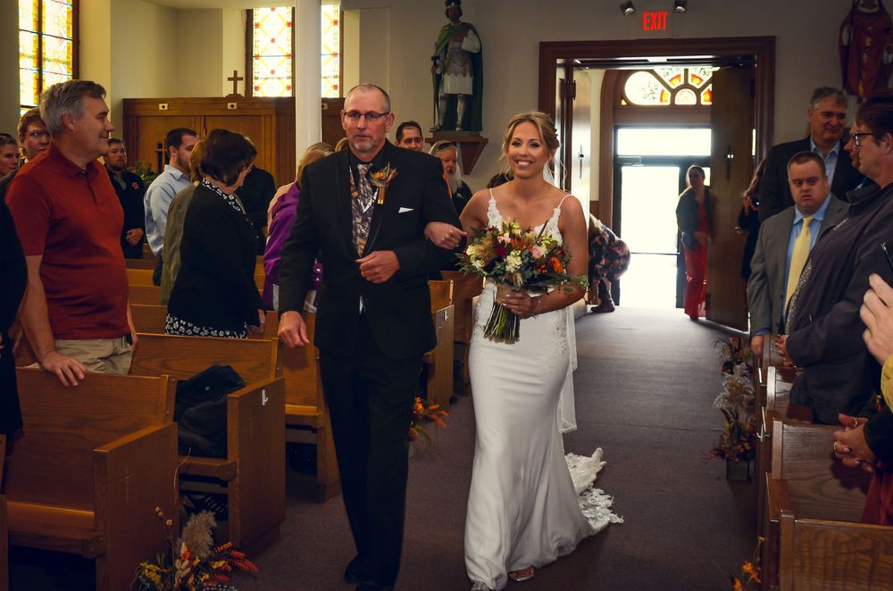A wedding procession walks down the aisle of a church with flowers and stained glass windows visible.