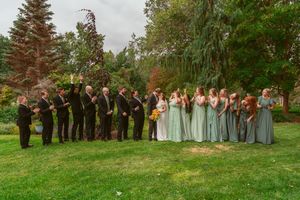 A wedding party in grey and sage green attire stands in a line on grass with evergreen trees in the background.