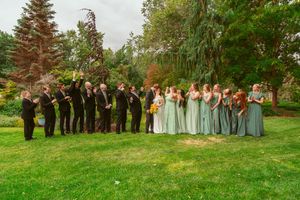 Wedding party wearing sage green and black attire stands together for outdoor group photos on grassy lawn near evergreen trees.