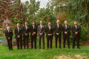 A group of gentlemen dressed in black suits and ties stand in line on green grass with lush gardens behind them.