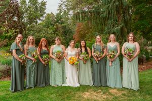 Group in sage green and mint dresses pose with floral bouquets on garden lawn.