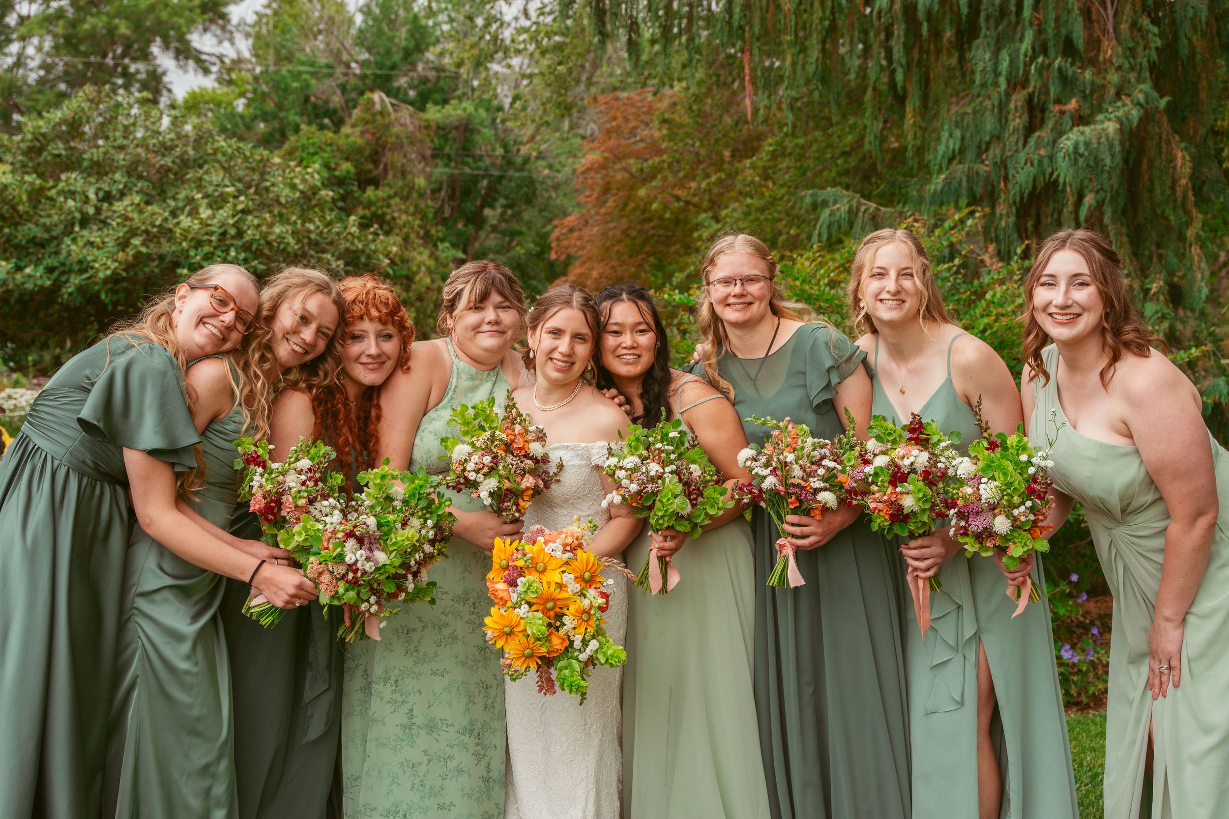 A group in coordinated sage and mint green gowns stands together holding vibrant autumn-colored floral bouquets.