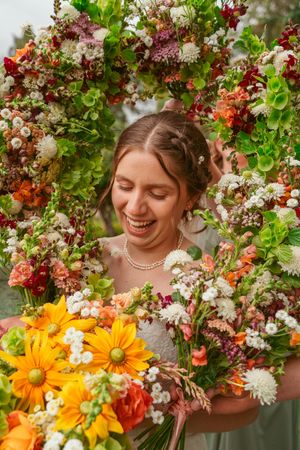 A person surrounded by colorful floral arrangements holds a bright bouquet of orange and white flowers while smiling.