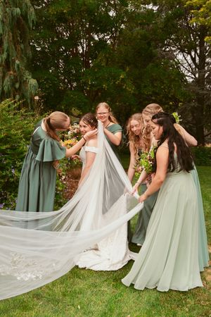 People in sage green dresses help arrange a flowing white wedding veil outdoors.