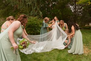 Bridesmaids in sage green dresses helping arrange a long bridal veil on grass.
