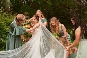 Bridesmaids in sage green dresses help arrange a flowing white wedding veil during an outdoor celebration.