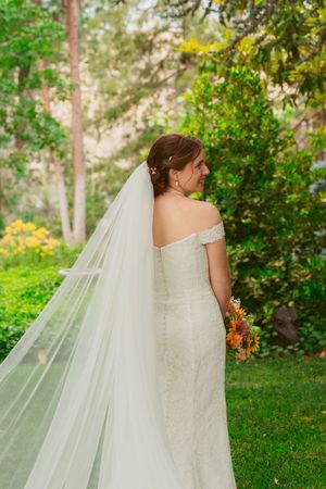 A bride in an off-shoulder white gown and flowing veil holds sunflowers while standing in a lush garden setting.