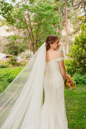 A bride in a white lace wedding dress poses among lush greenery with a flowing veil during an outdoor photoshoot.