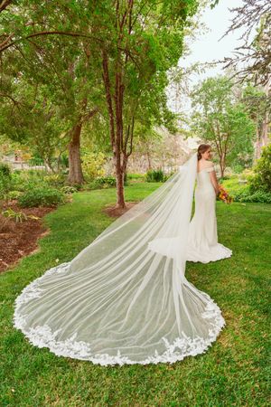 A stunning white wedding dress with an elaborate cathedral-length lace train spreads out on lush green grass.