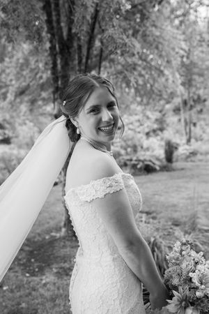 Black and white portrait of a bride in a lace wedding dress with a veil smiling outdoors in a garden setting.