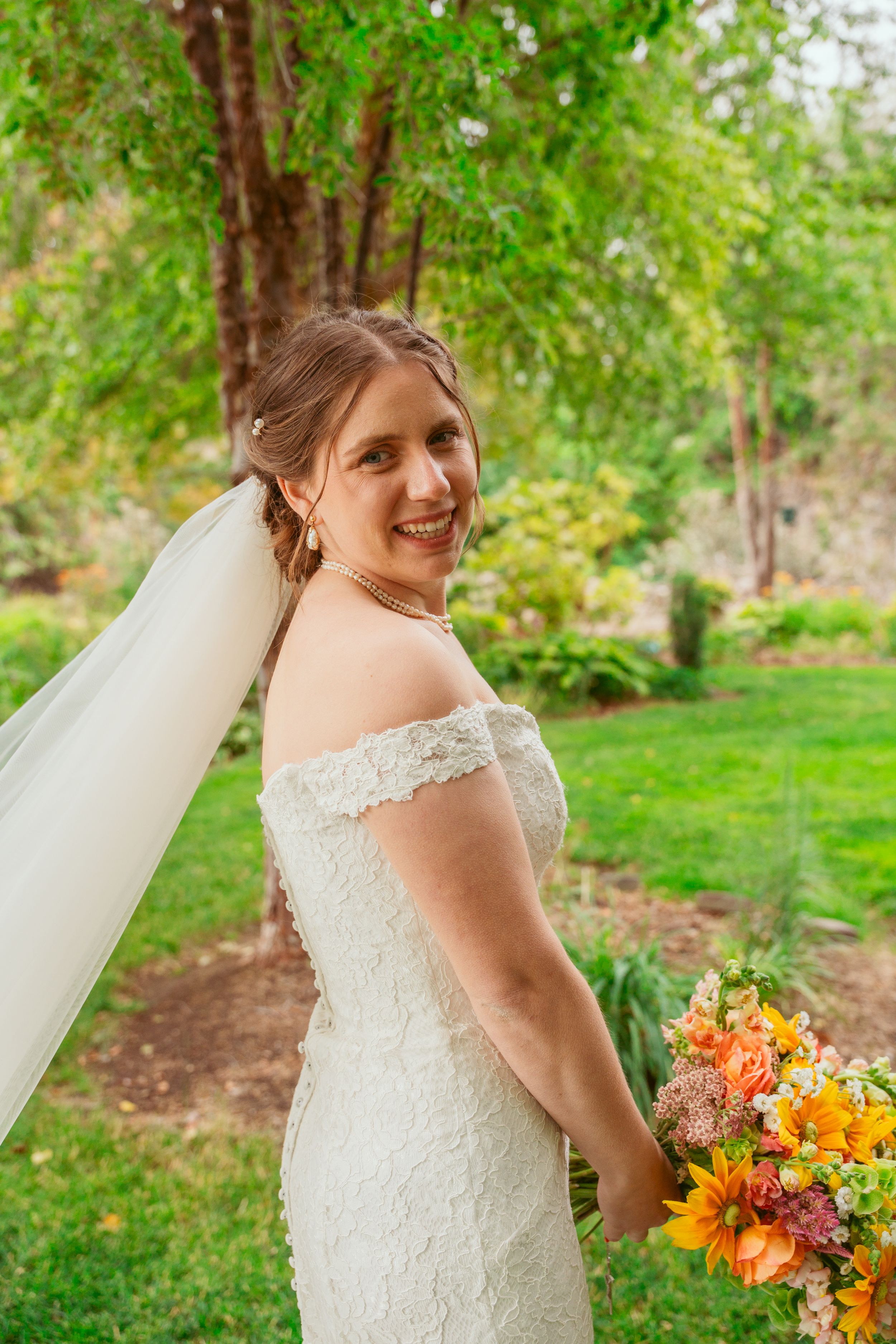 Bride in lace cap sleeve wedding gown holds vibrant orange and yellow bouquet in garden setting.