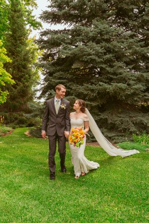 A couple walks through a garden path lined with evergreen trees on their wedding day.