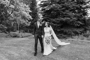 A black and white photo of a wedding couple posing outdoors in a garden with evergreen trees in the background.