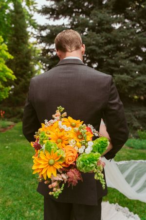 A person in a dark suit holds a vibrant bouquet with orange gerbera daisies and mixed flowers behind their back.