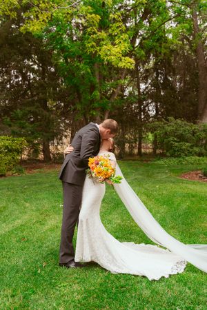Wedding couple embracing on grassy lawn with bride's long lace train and orange bouquet in autumn garden setting.
