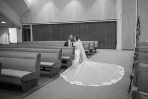 A black and white photograph captures a wedding moment in a church with long pews and a flowing cathedral-length veil.