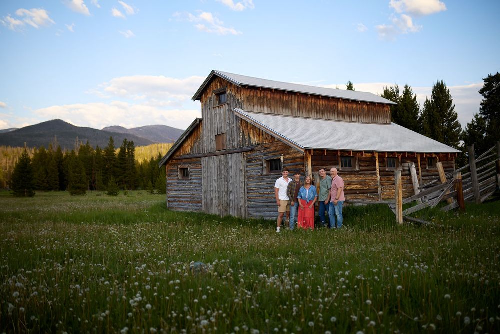 Rustic barn weathered by time stands against mountain backdrop as wildflowers bloom in grassy meadow at sunset.