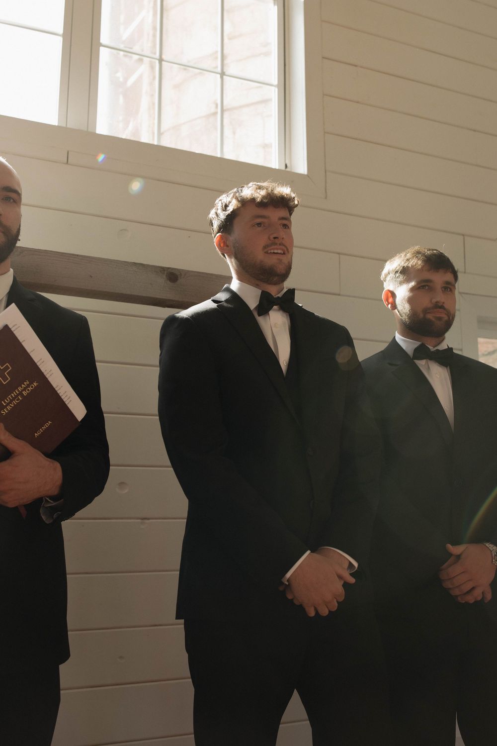 Groomsmen in black suits stand together in a bright indoor venue before a wedding ceremony.