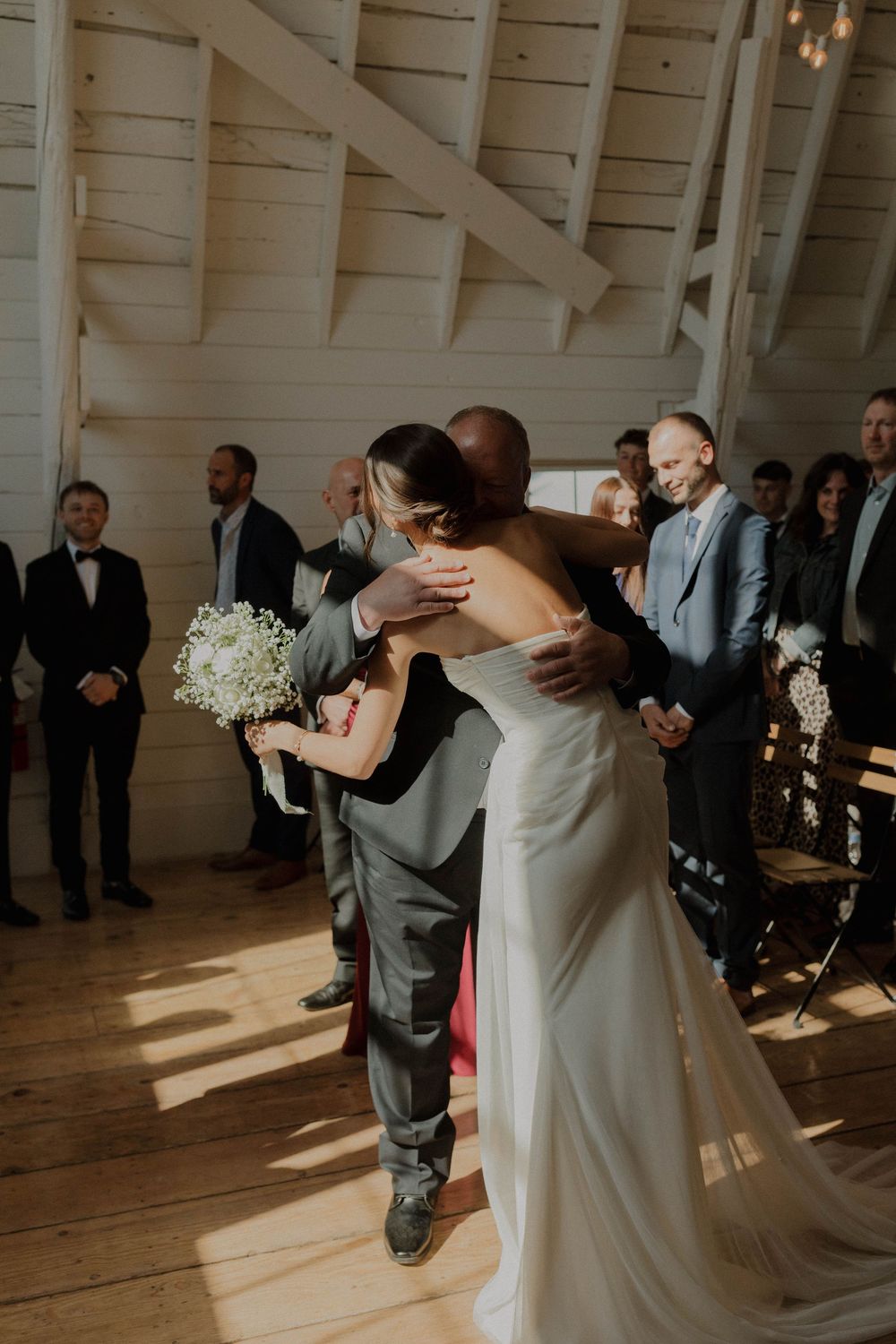 A couple shares their first dance in a rustic white-walled venue with natural light streaming through windows.