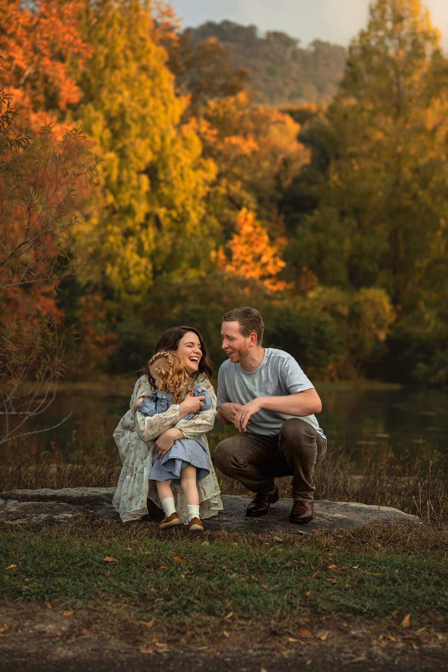 A family sits on a large rock surrounded by vibrant autumn foliage in a park setting during golden hour.