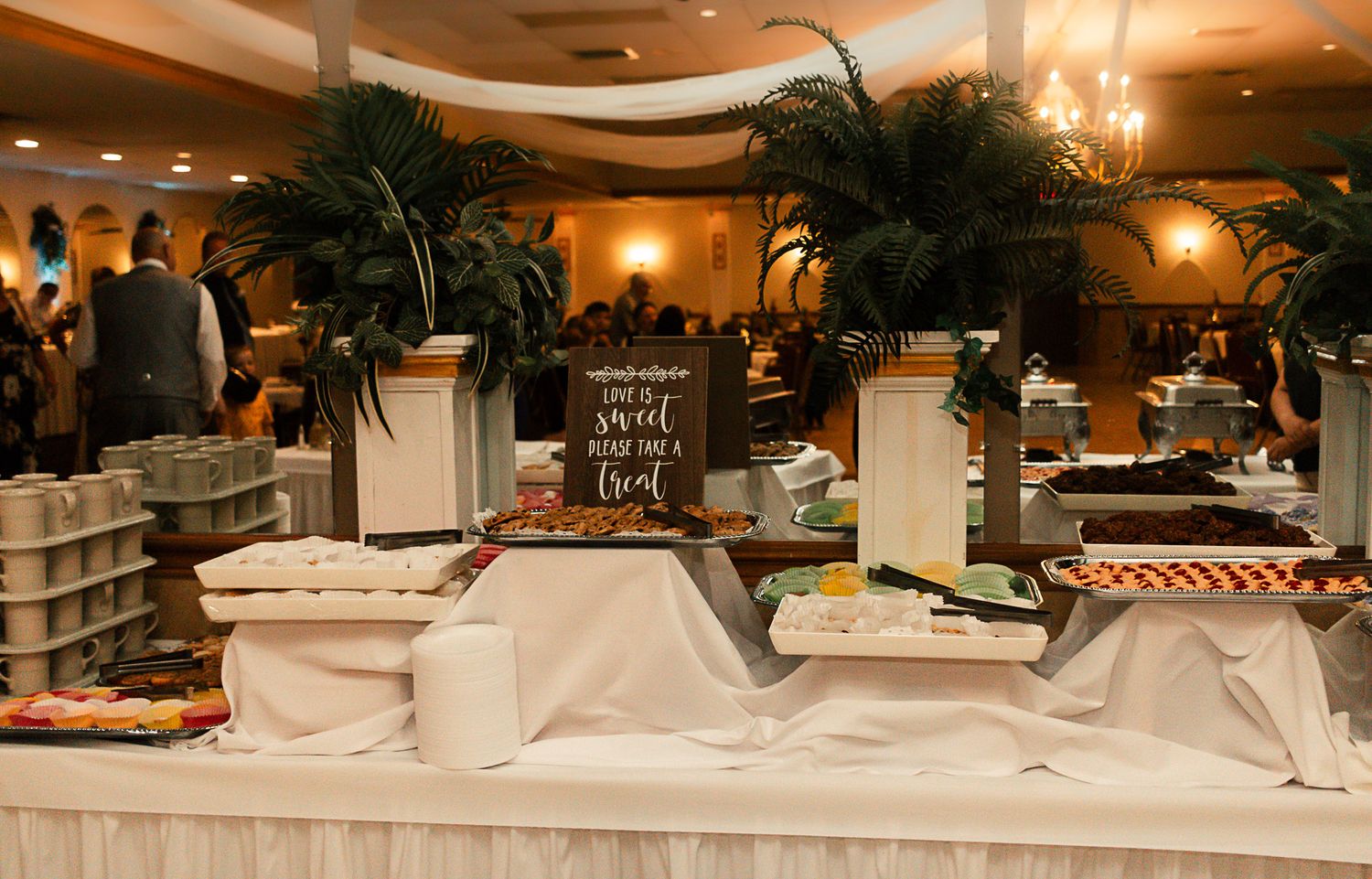 Elegant wedding banquet hall with white tablecloths and palm tree decorations under warm ambient lighting.