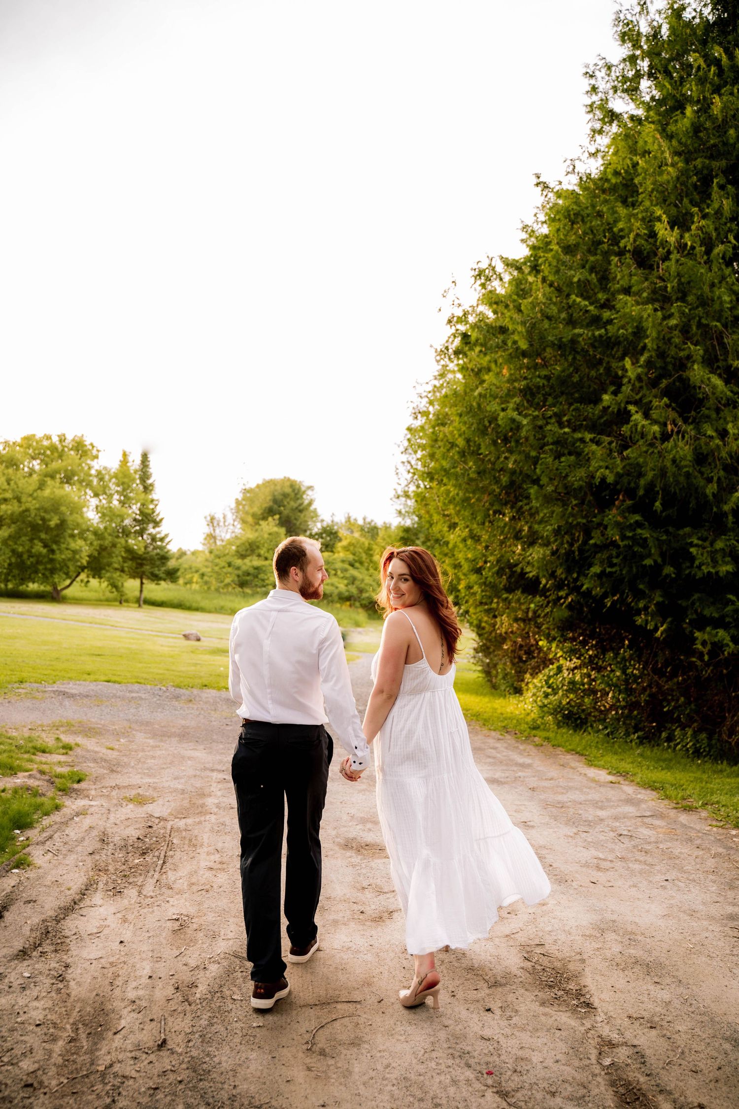 Couple in formal attire walking hand in hand down a dirt path surrounded by greenery during golden hour.