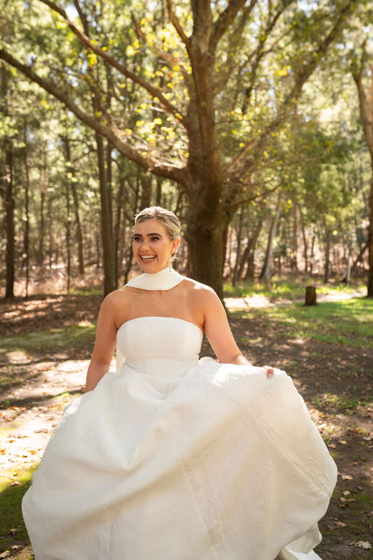 A bride in a strapless white wedding dress twirls joyfully in a sunlit forest setting.