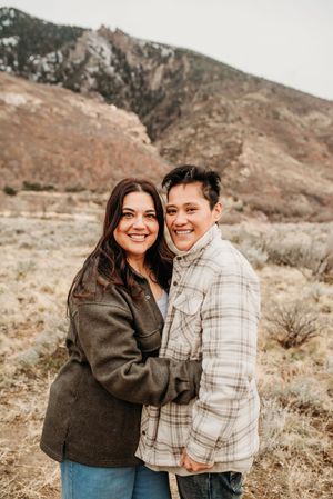 A couple embrace while standing in front of a rugged mountain landscape on a cloudy day.