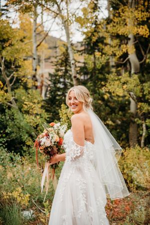 Beautiful autumn wedding portrait of bride in lace gown holding floral bouquet against golden foliage backdrop.