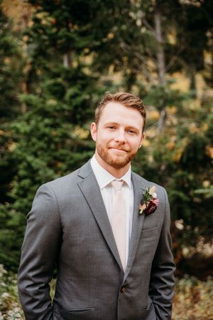 A groom in a grey suit and blush tie stands outdoors against a blurred forest background.