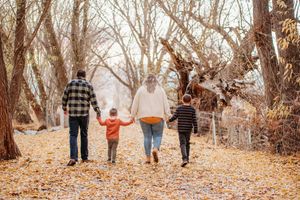 A group of people walk together holding hands along an autumn path lined with bare trees and fallen leaves.
