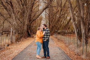 A couple stands on a leaf-covered path surrounded by towering autumn trees during a fall photo session.